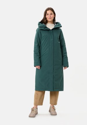 Donna in piedi che indossa un lungo cappotto invernale con cappuccio color verde teal, pantaloni beige corti e sneakers beige su sfondo bianco.