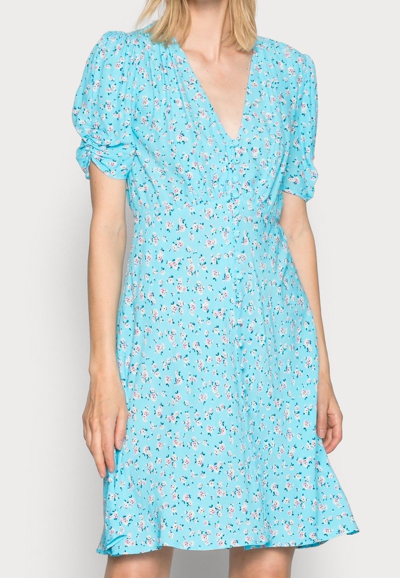 Femme portant une robe bleu clair avec un petit imprimé floral rose et blanc, manches bouffantes et encolure en V.