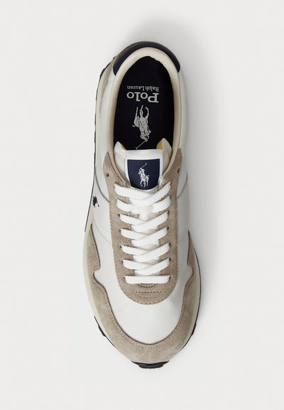 Balti un bēšu kedas ar baltām šņorēm, uz kurām ir Polo Ralph Lauren logo uz mēles un iekšpuses, redzamas no augšas uz baltas fona.