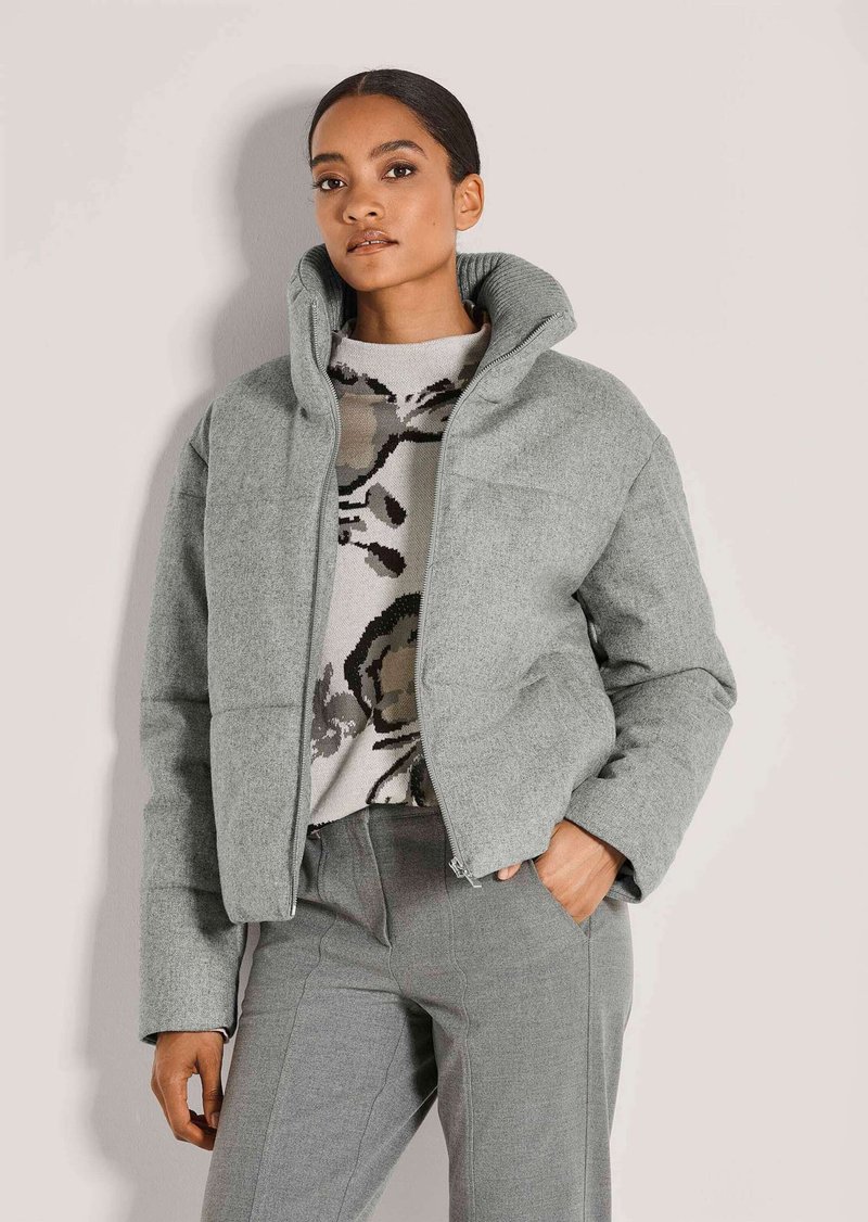 Frau trägt eine hellgraue Daunenjacke, einen Pullover mit Blumenmuster und passende graue Hose, steht mit einer Hand in der Tasche vor einem einfarbigen Hintergrund.
