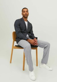 Grau kariertes Blazer, weißes Rundhals-T-Shirt, graue Jogginghosen und weiße Sneakers. Sitzt auf einem Holzstuhl mit schwarzem Sitz.