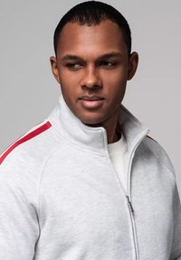 Giacca grigia con zip e collo alto, caratterizzata da strisce rosse sulle spalle e una texture liscia, indossata sopra una camicia bianca.