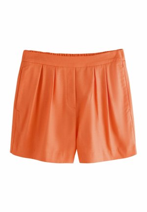 Frau mit langen Haaren, die eine orange ärmellose, geknöpfte Weste und passende hoch taillierte Shorts trägt und an einem Fenster mit Blick auf eine Küstenlandschaft steht.