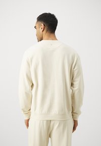 Cream-colored sweatshirt with a crew neck, long sleeves, and ribbed cuffs, featuring a small circular detail at the back neckline.
