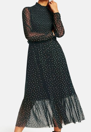 Vestido largo negro con cuello alto y mangas largas transparentes. Presenta un estampado de puntos y un dobladillo con volantes en capas, confeccionado con tela ligera.