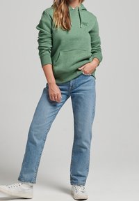 Felpa verde con tasca frontale, cappuccio regolabile, abbinata a jeans dritti azzurri e sneaker bianche, su uno sfondo neutro.