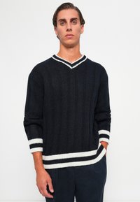 Pull en tricot marine avec un col en V rayé blanc et des poignets. Présente une texture côtelée et une coupe décontractée avec des manches longues.