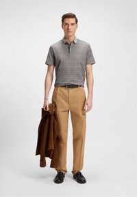 Hombre con una camisa de polo gris con patrón y pantalones color beige, sosteniendo una chaqueta marrón, de pie sobre un fondo blanco liso.