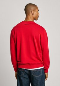 Sudadera roja de algodón con cuello redondo, mangas largas, puños y dobladillo acanalados. Presenta un logo sutil en la parte posterior.