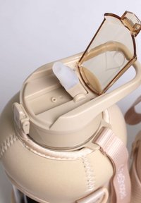 Bouteille d'eau beige avec un couvercle rabattable clair et des accents marron. Présente un bec en silicone, une poignée intégrée et une housse en tissu avec des coutures.