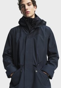 Marineblå vattentät jacka med zippered fram, justerbar huva och två framfickor. Slät textur och minimalistisk design, med tryckknappar.