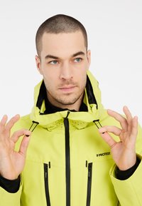 Veste de ski jaune avec une capuche, cordons de serrage réglables, zips noirs et aérations. Logo de la marque sur la zone de la poitrine.