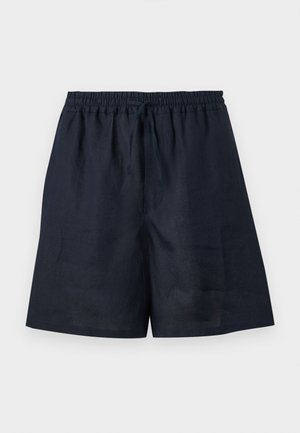 Joven con camisa de manga corta a rayas, pantalones cortos azul marino, sandalias negras, sosteniendo una bolsa negra con cordón, de pie sobre un fondo blanco liso.