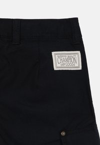 Juodos audinio šortai su siuvinėtu užtrauktuku, pažymėtu "Champion Dry Goods". Yra standartinės diržo kilpos ir mygtukas prie apatinio krašto.