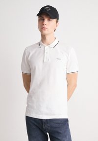 Witte poloshirt met marineblauwe kraagstrepen en logo op de borst, gedragen met donkere jeans en een zwarte pet. Soepel katoenmateriaal.