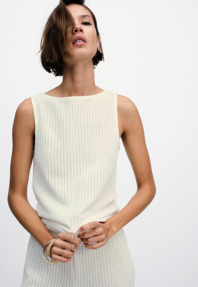 Žena s krátkými hnědými vlasy má na sobě krémový pletený top bez rukávů a sukni, upravuje látku v pase, na jednobarevném bílém pozadí.