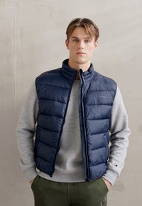 Navy-blauer Steppwesten mit Reißverschluss, versehen mit horizontalen Steppnähten. Getragen über einem hellgrauen Sweatshirt und olivgrünen Hosen.
