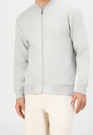 Mann trägt eine hellgraue Jacke mit Reißverschluss und cremefarbene Hose und steht vor einem einfarbigen hellen Hintergrund.