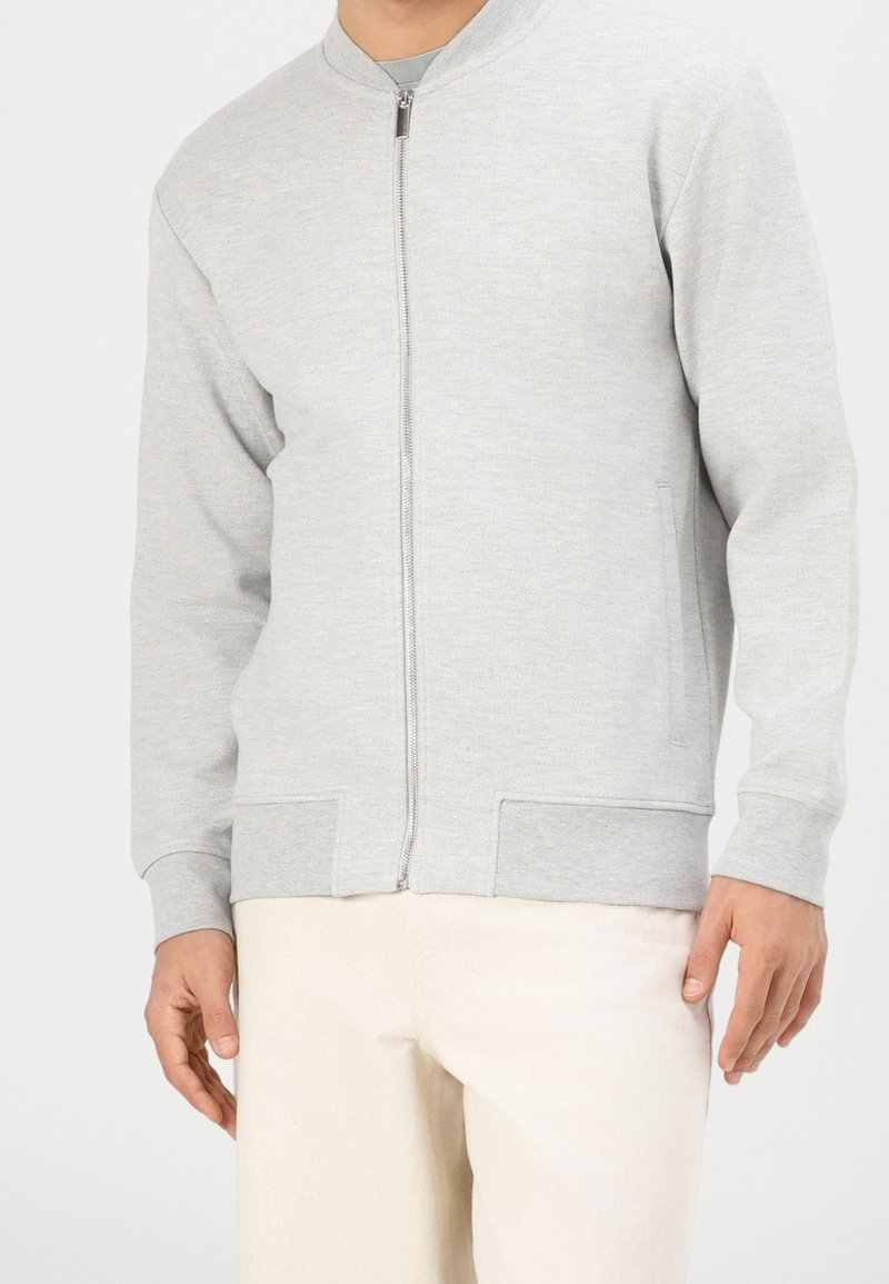 Homme portant une veste zippée gris clair et un pantalon blanc cassé, debout devant un fond clair uni.