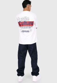 White t-shirt with bold red and blue typography on the back, stating "Hustle Culture." Paired with navy cargo pants and white sneakers.