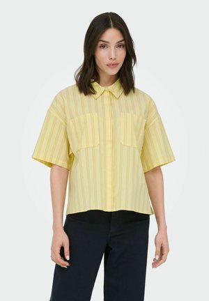 Femme portant une chemise jaune à manches courtes avec des rayures verticales et un pantalon noir, debout devant un fond clair uni.