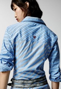 Cămașă albastră cu dungi, cu mâneci suflate, având linii orizontale și verticale, și un mic logo roșu brodat pe spate.