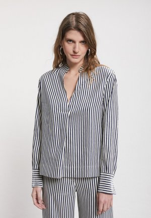 Woman wearing matching navy and white vertical striped blouse and pants standing against plain background facing forward.