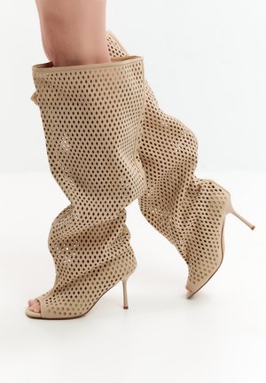 Beige kniehohe Stiletto-Stiefel mit diamantförmigen Perforationen und offenen Zehen, getragen auf nackten Füßen.