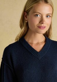 Marineblauer V-Ausschnitt-Pullover mit gerippter Textur, lockerer Passform und überschnittenen Schultern. Eine goldene Halskette und Ohrstecker aus Ringen werden als Akzente getragen.