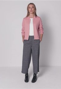 Veste zippée rose clair portée sur un haut blanc, associée à un pantalon gris large et texturé ainsi qu'à des bottines noires. Design minimaliste.