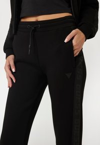 Black sweatpants with a smooth texture, elastic waistband, and a drawstring. Features a logo patch and a printed brand name on the side.