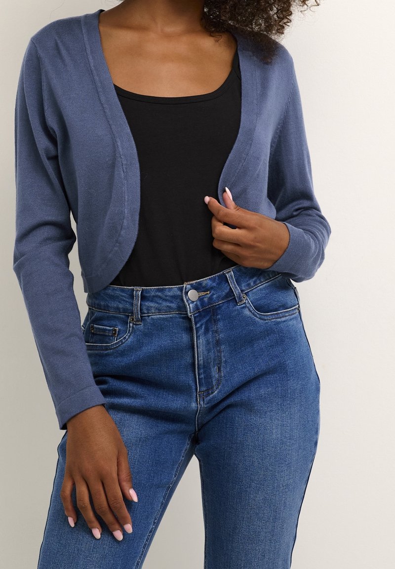 Femme portant un cardigan bleu court sur un haut noir et un jean taille haute bleu, debout devant un fond clair uni.
