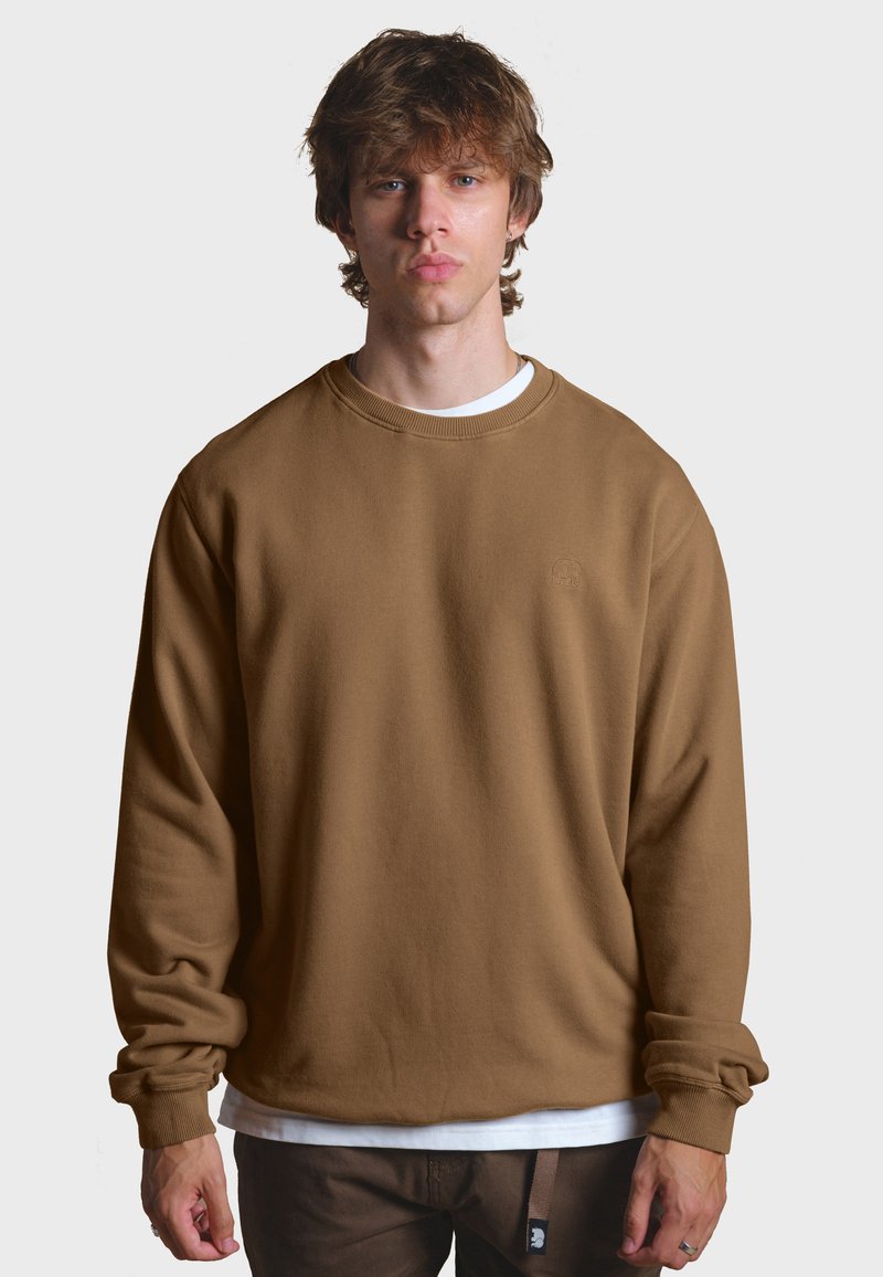 Brauner Pullover mit Rundhalsausschnitt, langen Ärmeln, gerippten Bündchen und dezentem Logodetail auf der Brust. Getragen über einem weißen T-Shirt.
