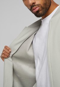 Giacca zip grigia chiara con una texture liscia, che mostra un interno morbido. Indossata sopra una t-shirt bianca a girocollo, per un look informale.