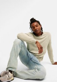 Lichtgrijs overhemd met lange mouwen, relaxte pasvorm, denim jeans, en grijze sneakers. Model zit op een witte achtergrond in een casual houding.