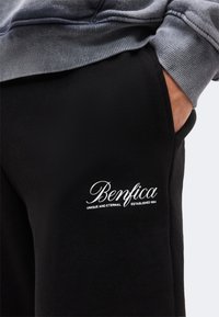 Άτομο που φοράει μαύρο παντελόνι με το κείμενο «Benfica Unique and Eternal Established 1904» και γκρι φούτερ, χέρι στην τσέπη.