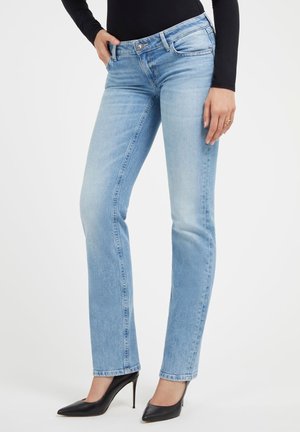 Kvinde iført lyseblå jeans med lige ben og sorte højhælede sko, hånd hviler i lommen, mod ensfarvet baggrund.