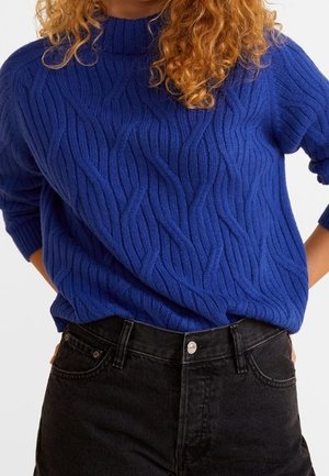Persona che indossa un maglione blu a trecce infilato in jeans neri a vita alta con le mani posate sui fianchi.