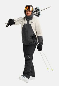 Skiausrüstung mit einer weißen und grauen Jacke mit Reißverschlüssen, schwarzen Hosen, schwarzen Handschuhen und Skistöcken, vor einem schlichten Hintergrund.