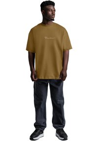 Maglietta marrone oversize con maniche corte e scollo rotondo, con testo bianco in corsivo sulla parte anteriore. Abbinata a pantaloni scuri larghi.