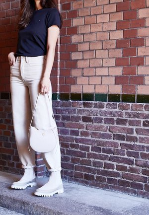 Femme debout contre un mur en briques, portant un t-shirt noir, un pantalon crème, des bottes beige clair et tenant un petit sac à main beige.