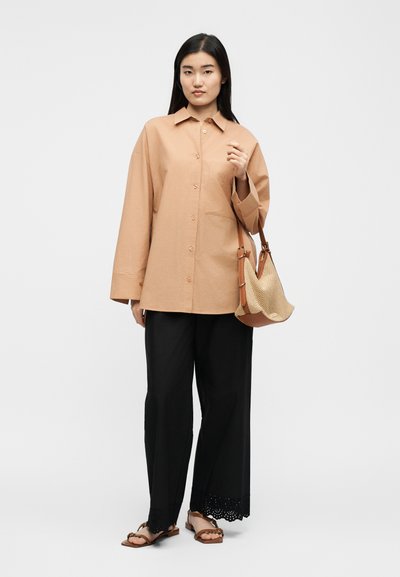 Γυναίκα με άνετο μπεζ πουκάμισο με κουμπιά, μαύρο παντελόνι φαρδιάς γραμμής με δαντελωτό τελείωμα, καφέ σανδάλια και κρατά μια μπεζ υφαντή τσάντα στον ώμο της.