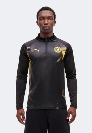 Man draagt een zwarte sporttop met lange mouwen met een geel Puma-logo en Borussia Dortmund BVB 09 embleem, staand tegen een effen lichte achtergrond.