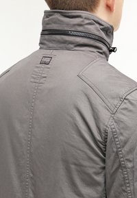 Szara kurtka z wysokim kołnierzem, wyposażona w czarny zamek błyskawiczny i małą metkę z logo marki na plecach. Wykonana z gładkiego, teksturowanego materiału.