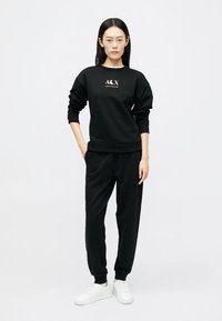 Melni sporta bikses un džemperis, kas izgatavoti no mīksta auduma, ar balto logo uz krūtīm, komplektā ar baltām sporta apaviem.