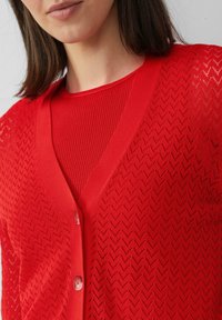 Roter Cardigan mit V-Ausschnitt und Knopfverschluss, der ein Zickzackmuster aufweist und aus leichtem, strukturiertem Stoff besteht, der über einem gerippten roten Oberteil getragen wird.