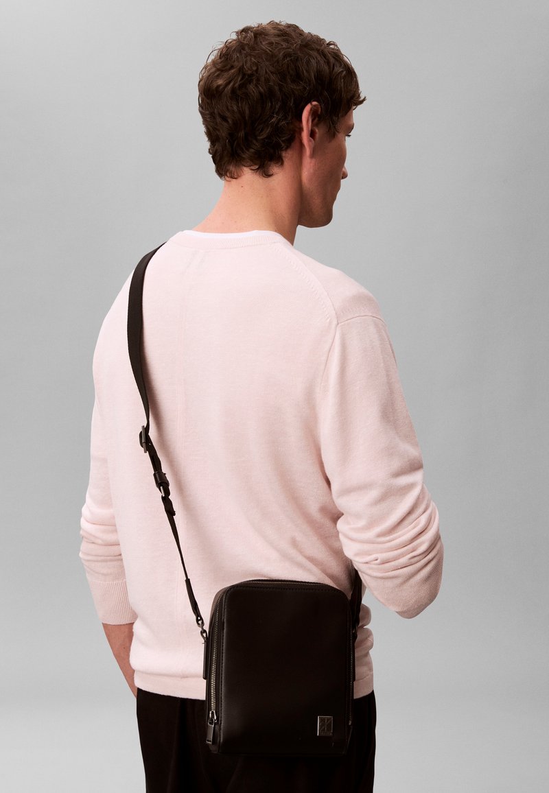 Homme portant un pull rose pâle et un sac bandoulière noir avec une bandoulière ajustable, tourné de dos sur un fond gris uni.