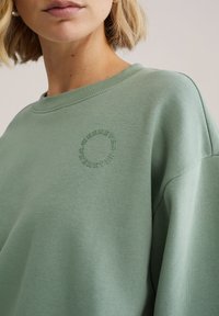 Lichtgroene trui met een rond geborduurd logo met de tekst "WHEREVER" langs de zijkant. Zacht, gevoelig materiaal en een ontspannen pasvorm.