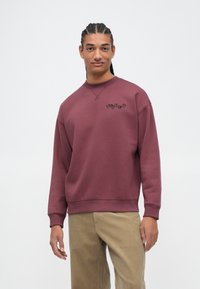Sweatshirt bordeaux avec un col rond et un logo noir sur la poitrine, fabriqué en coton. Comprend des poignets et un ourlet côtelés, coupe décontractée.
