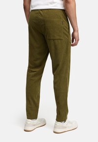 Uomo con pantaloni in velluto a coste verde oliva con tasca posteriore e sneakers bianche, in piedi contro uno sfondo semplice.